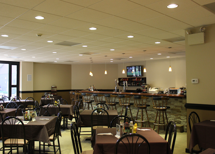 Bar and Grille. The foreground of the image shows the dining tables, primarily set for four people. 10 barstools are positioned at the bar. A television is on the wall on the left side of the bar.