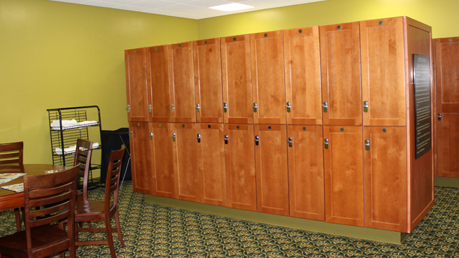 The men's locker room at Buena Vista Country Club. There are two rows of half-size and full-size wood-stain-finished lockers in the room. Chairs are set at a round card table, and a towel rack is also in the room.