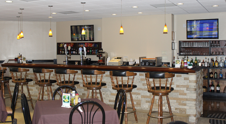 Bar and Grille. Table setting for four, set with a linen, utensils, and condiments are in the foreground. Several barstolls line the bar. Two television are turned on hanging on the wall behind the bar.