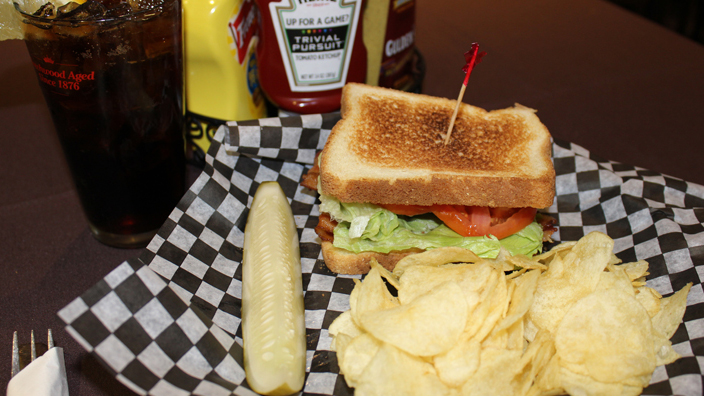 A B.L.T. Bacon, lettuce, and tomato on toasted white bread with a pickle and potato chips. A glass of diet coke and condiments are behind the serving dish.