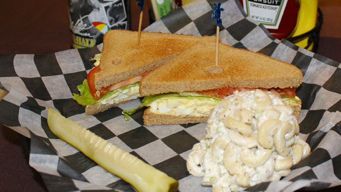 An Egg Salad Sandwich. Egg salad topped with lettuce and tomato on toasted wheat bread. On the side, are a pickle and macaroni salad. A bottle of Arnold Palmer is served with the sandwich.
