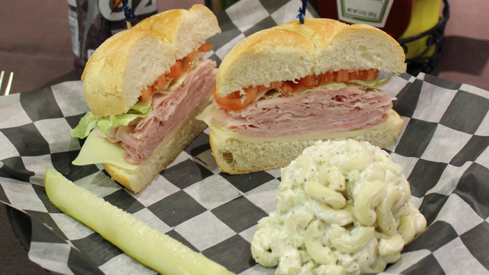 A Ham & Cheese Sandwich. Stack of ham placed over top cheese topped with lettuce and tomato on a kaiser roll cut in half. Sandwich is served with macaroni salad and a pickle.