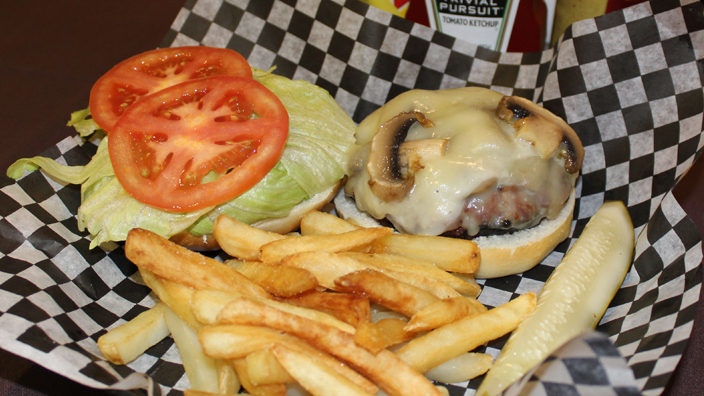 The Bunker Swiss Shroom special menu item. A burger patty covered in melted swiss cheese with sliced mushrooms in and on the cheese. The burger is on the bottom half of a pub roll while the top half of the roll is placed beside it with lettuce and tomato. French fries and a pickle spear are on the side.