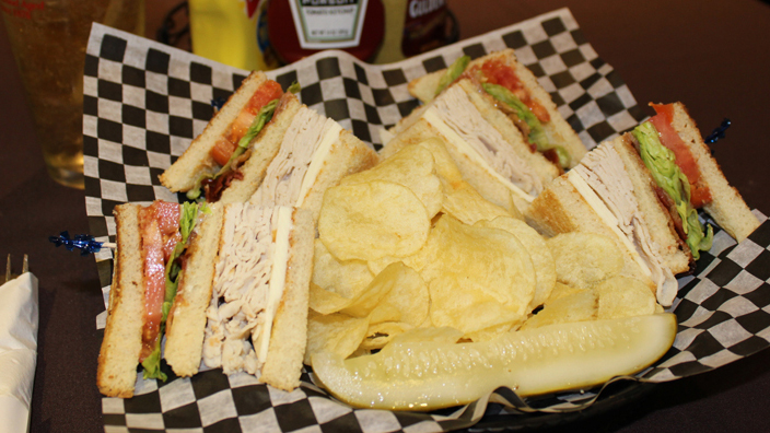 A turkey club. The plate is full with four turkey club Wedges each with a toothpick holding the three slices of bread, turkey, bacon, cheese, lettuce and tomato together. The rest of the plate has potato chips and a pickle spear.