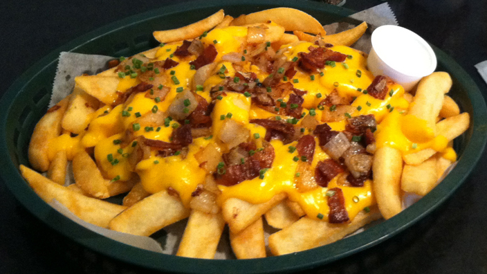 Fatty Fries special menu item. In a round, plastic food basket, thick-cut steak fries are topped with smooth and melted cheddar cheese, diced, caramelized onions, and bacon pieces. A ramekin of sour cream is on the side.