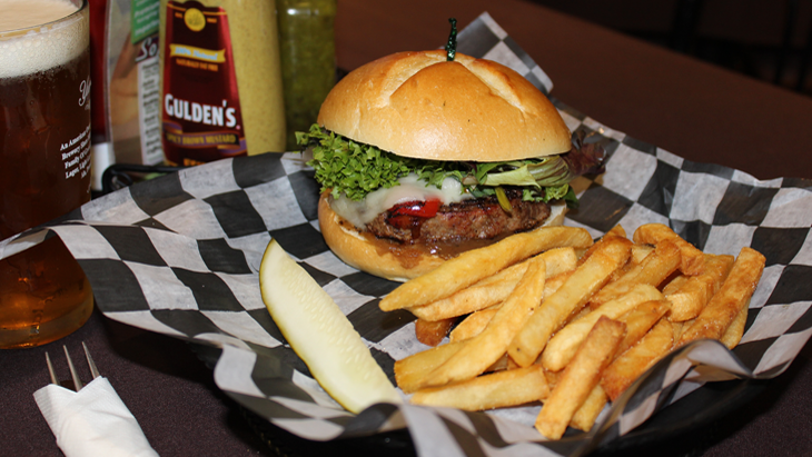 The Little Italy special menu item. Placed in a pub roll, a burger patty is flavored with Balsamic-dressed spring-mix greens and topped with melted provolone cheese and roasted red peppers. French fries and a pickle spear are on the side. A glass of beer and condiments are in the background.