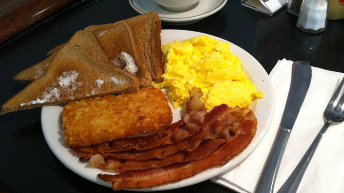 Breakfast platter served on a white dinner plate. On the plate are four half-slices of buttered wheat bread fanned out. In addition, there are scrambled eggs, four slices of bacon, and a fried hashbrown. A coffee cup is in the background and silverware is placed next to the plate.