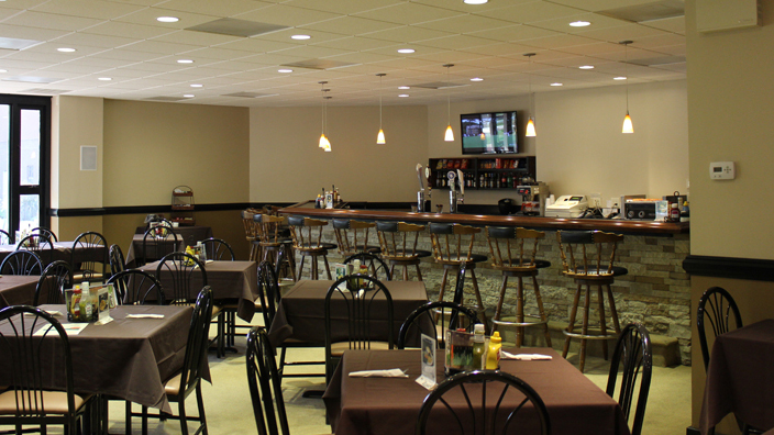 Bar and Grille. The foreground of the image shows the dining tables, primarily set for four people. 10 barstools are positioned at the bar. A television is on the wall on the left side of the bar.