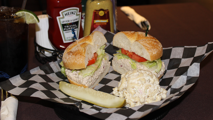Tuna salad, lettuce and tomato on a kaiser roll cut in half. On the side are macaroni salad and a pickle spear. In the background, there is a ketchup bottle, spicy mustard, and a glass of soda.